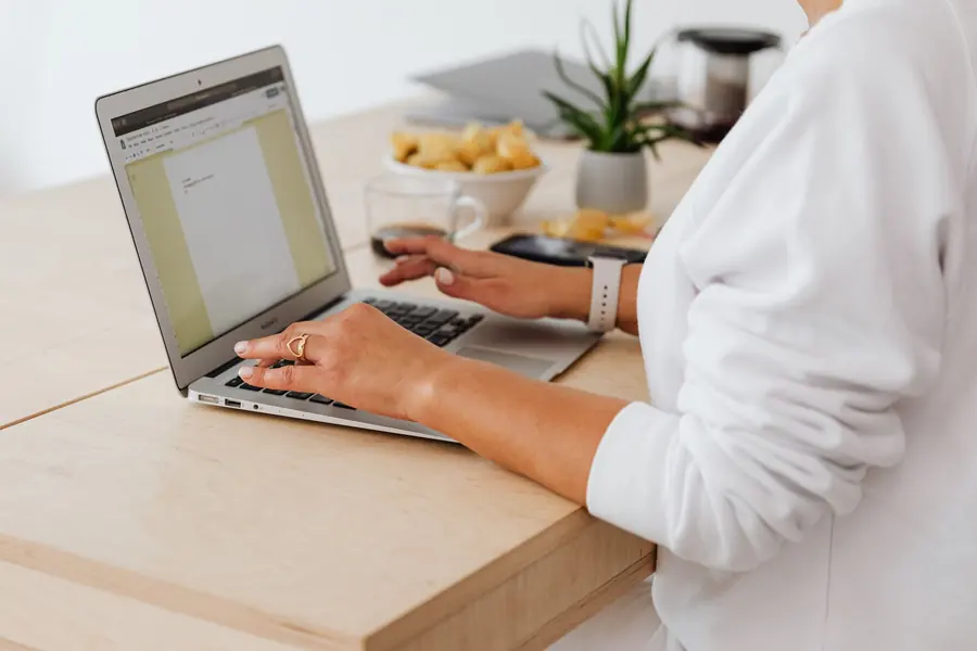 A person at a home desk reviewing paperwork on a laptop beside a notebook.