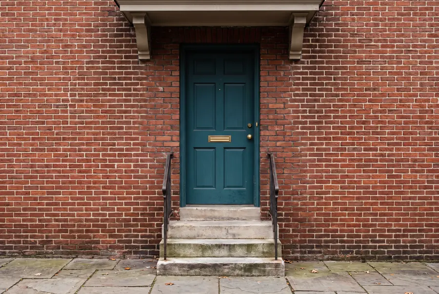 The teal front door of a 1920s American brick rowhouse with brass fittings and three concrete steps.